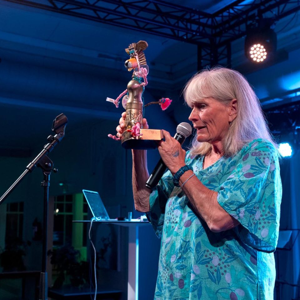 Antonia Ringbom, dressed in a blue dress, speaks into a microphone while holding a self-made award.