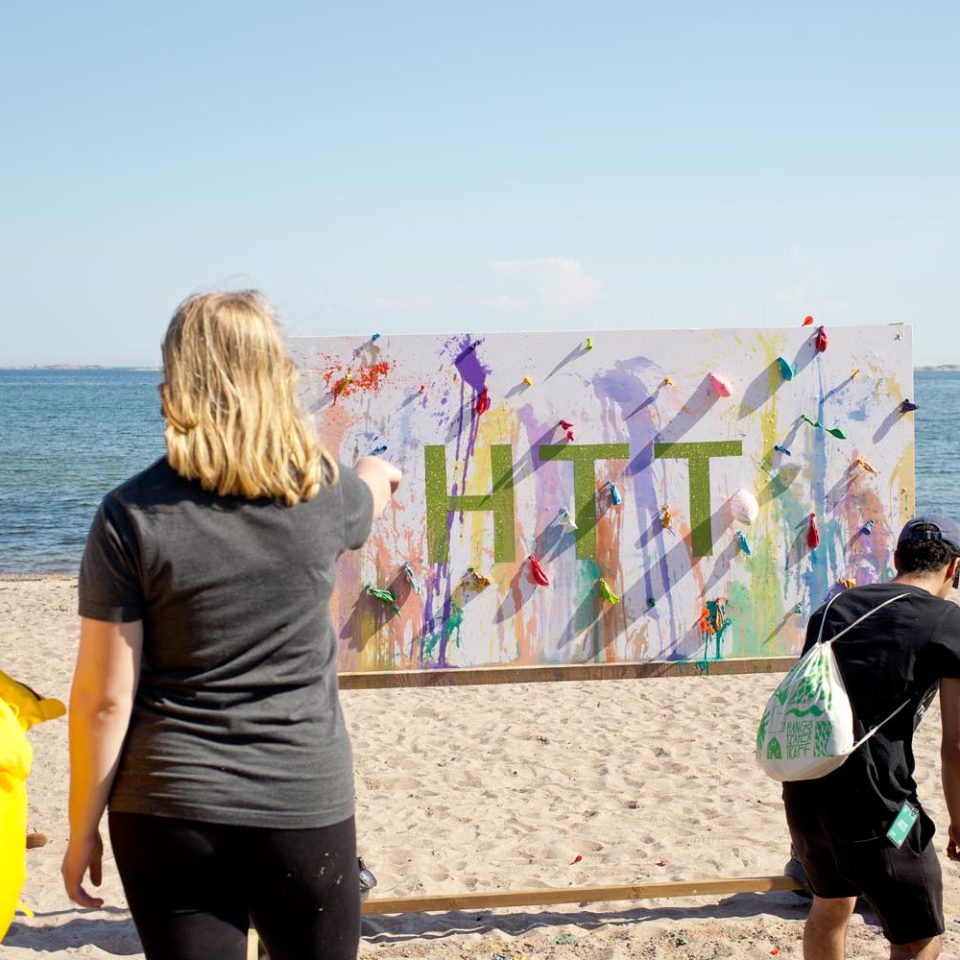 Four youths stand with their backs to the camera, one throwing a dart at a big panel that says HTT and on which balloons filled with paint have been popped. The ocean lies in the background.