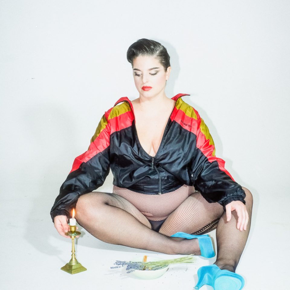 Ann-Sofie Lundin sits on a white floor looking at a candle, wearing fishnet stockings, blue high heels, and a shell suit jacket.