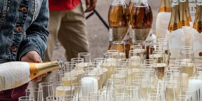 Många champagneglas på ett bord med Pommac- och skumpaflaskor i bakgrunden. En person håller på att hälla skumpa i glasen.
