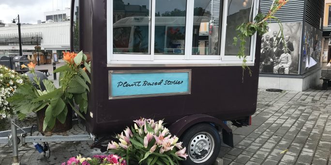En vagn med tak som befinner sig mitt på en torg; vagnen omringas av blommor och den har en skylt där det står "Plant Based Stories".