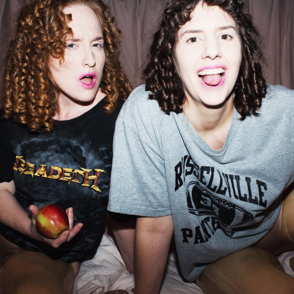 Nadja Hjorton och Lisen Rosell klädda i stora t-skjortor, sitter på en säng och tittar mot kameran. Ena har ett äpple i handen.