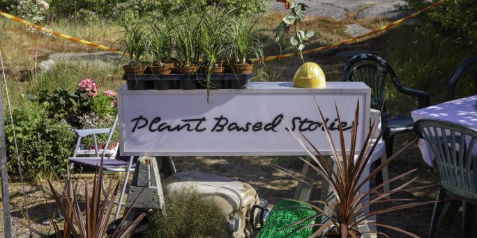 En skylt där det står Plant Based Stories ligger ute, omringad av olika växtligheter.
