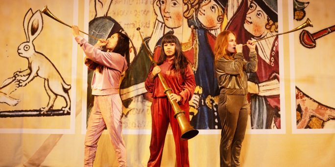 Anna Vnuk, Sandra Medina och Lisen Rosell står med trumpeter i handen, klädda i velour. I bakgrunden syns medeltida bilder.