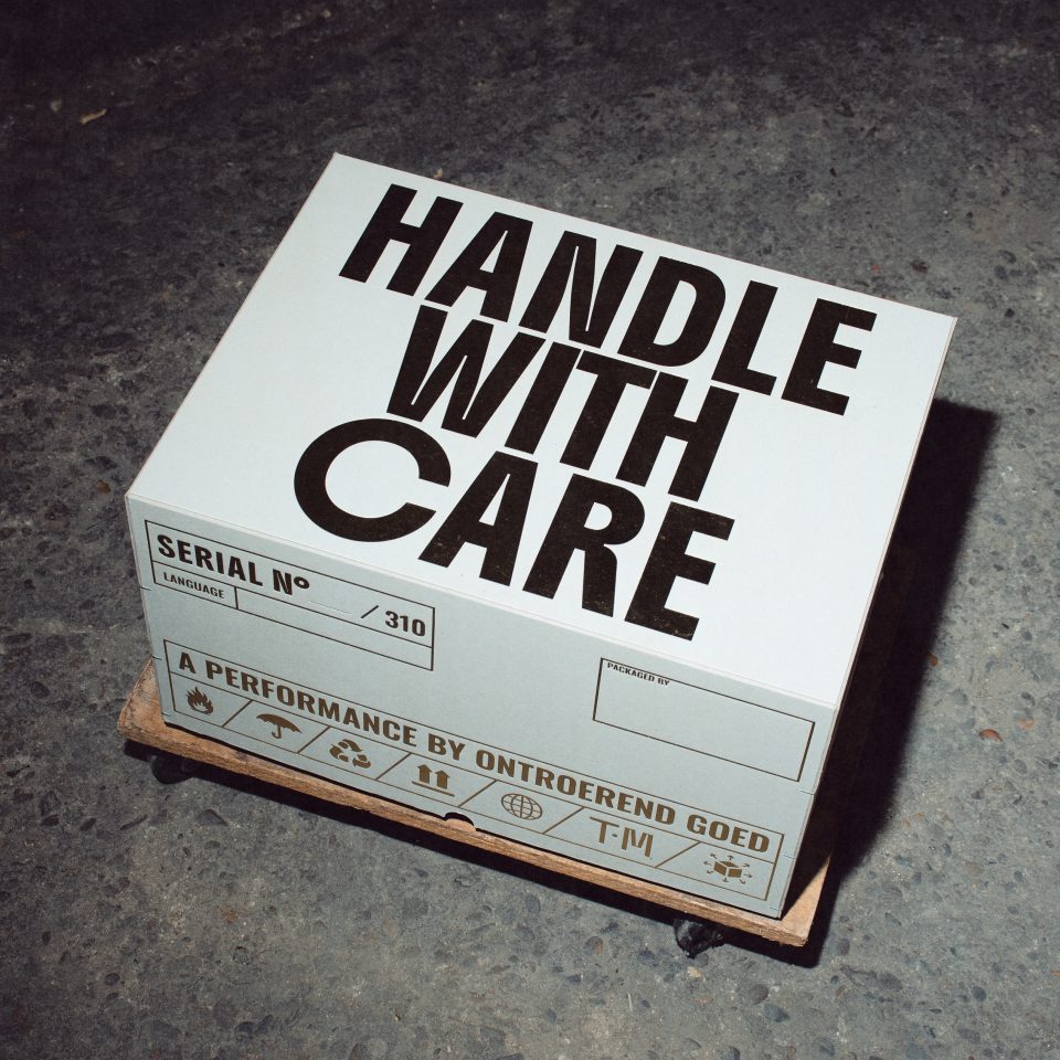 På golvet ligger en låda där det står med stor text "Handle with Care".