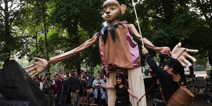 En stor docka i parken, med en svartklädd konstnär framför dockan och publiken bakom den.
