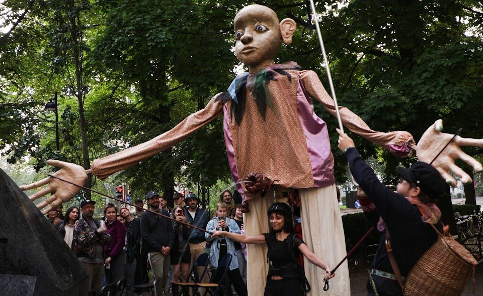 En stor docka i parken, med en svartklädd konstnär framför dockan och publiken bakom den.