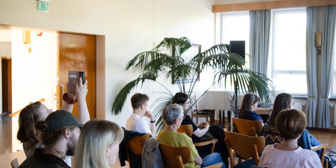 Publiken sitter på stolar i stadshusets foajé och lyssnar på en föreläsning.